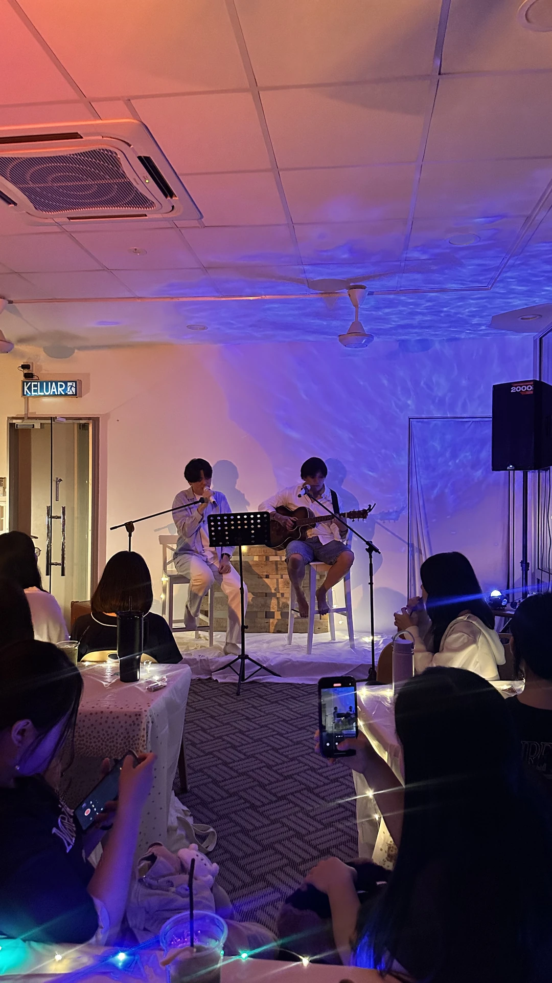 2026年7月的小型音乐会，两位演唱者在蓝紫灯光下弹唱，观众围桌而坐